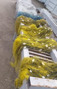 Yellow fishing nets with white floats draped over a white wooden bench on a boat dock area, blue nets visible in the background