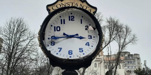 Public street clock with a Constanta sign, weathered face and blue numerals, broken glass, set in a square with bare trees in the background