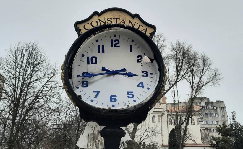 Public street clock with a Constanta sign, weathered face and blue numerals, broken glass, set in a square with bare trees in the background