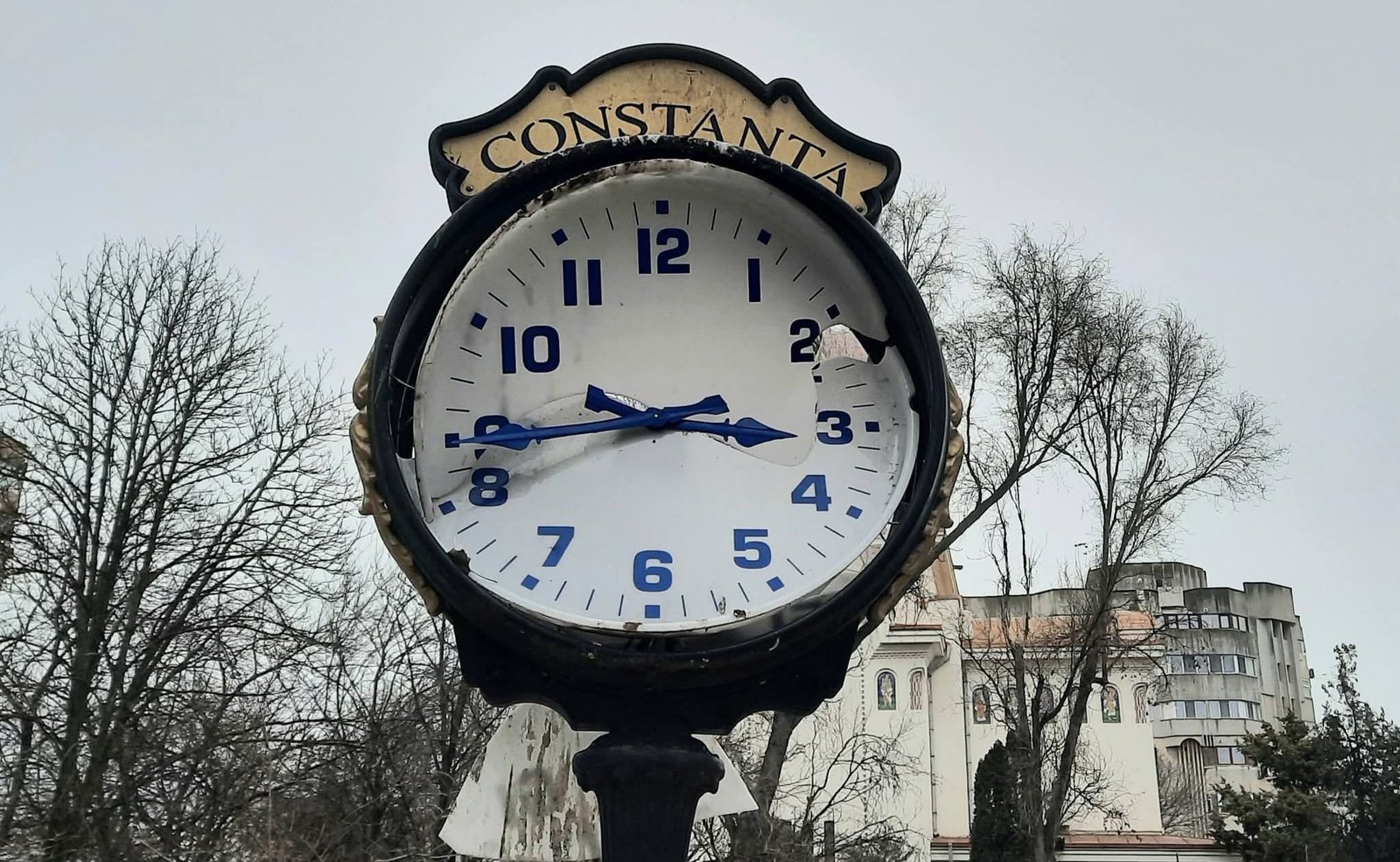Public street clock with a Constanta sign, weathered face and blue numerals, broken glass, set in a square with bare trees in the background