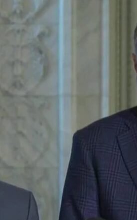 Two men in suits standing indoors during a press briefing, looking serious.