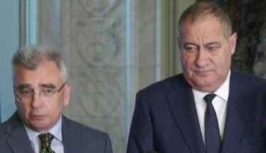 Two men in suits standing indoors during a press briefing, looking serious.
