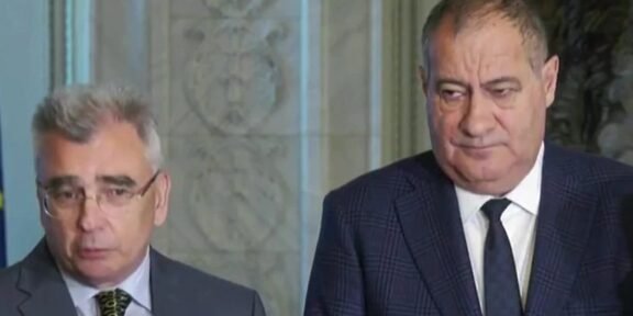 Two men in suits standing indoors during a press briefing, looking serious.