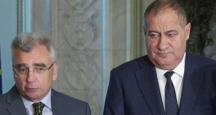 Two men in suits standing indoors during a press briefing, looking serious.