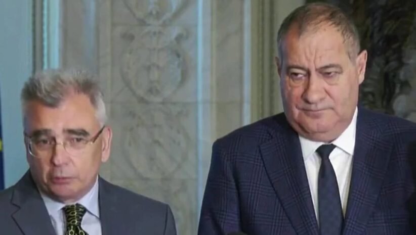 Two men in suits standing indoors during a press briefing, looking serious.