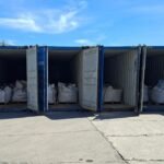 Four open blue shipping containers on a concrete lot, each filled with large white bags on wooden pallets under a sunny sky.