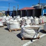 Industrial yard with large white bulk bags on pallets, blue shipping containers, and workers in high-visibility vests in the background, under a clear sky.