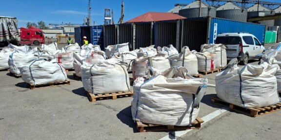 Industrial yard with large white bulk bags on pallets, blue shipping containers, and workers in high-visibility vests in the background, under a clear sky.
