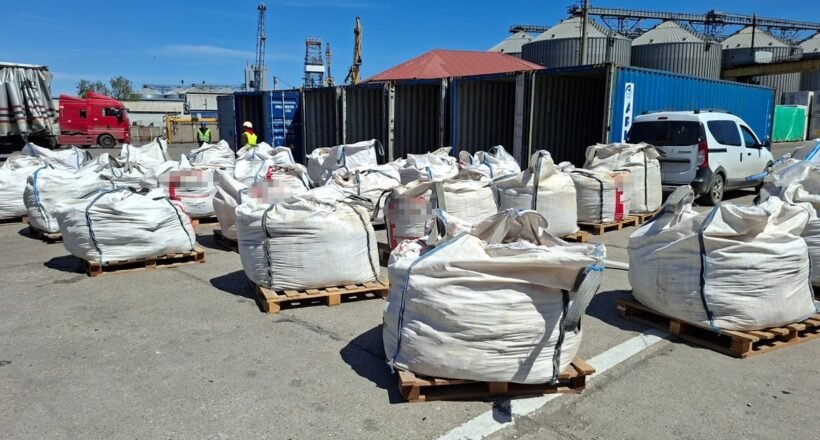Industrial yard with large white bulk bags on pallets, blue shipping containers, and workers in high-visibility vests in the background, under a clear sky.