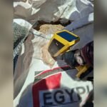Handheld yellow testing device with a blue screen held by a gloved hand over a soil-filled bag at a field site, used for on-site analysis
