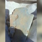 Rusty, rust-brown metal plate fragment with chipped edges and dirt on its surface, placed on a light fabric background.