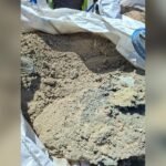 Excavation site showing loose soil in a trench with a white tarp nearby and safety gear at the top edge of the photo.