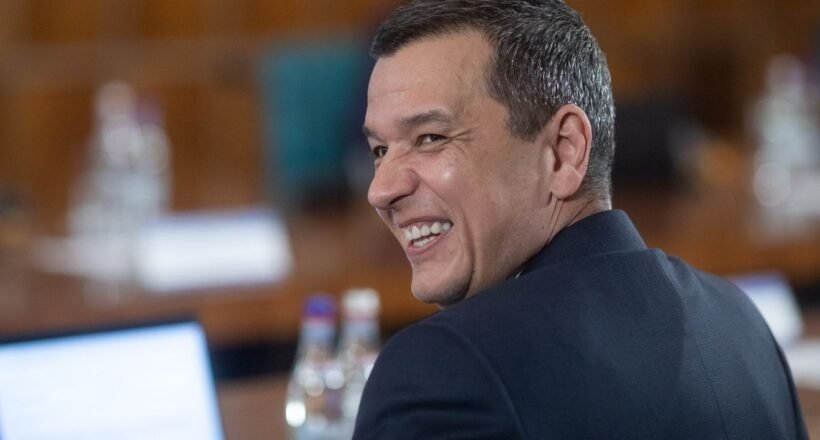 Man in a dark suit smiling over his shoulder in a conference room at a table with water bottles nearby
