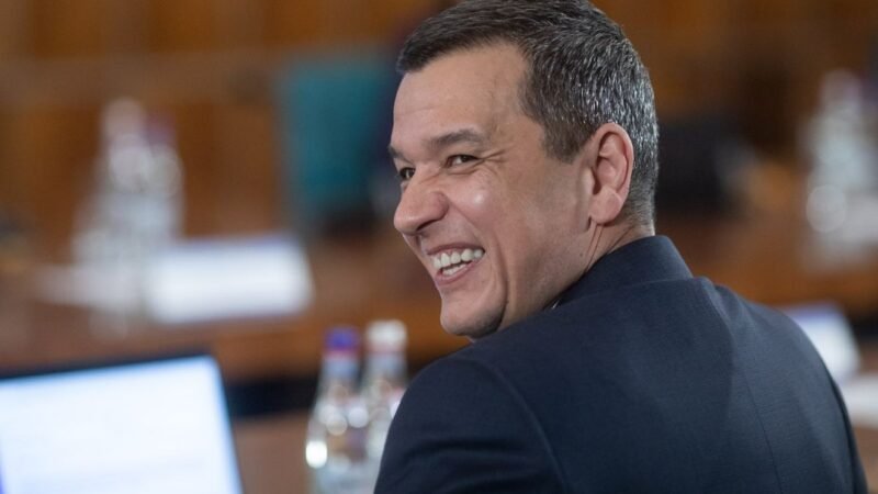 Man in a dark suit smiling over his shoulder in a conference room at a table with water bottles nearby