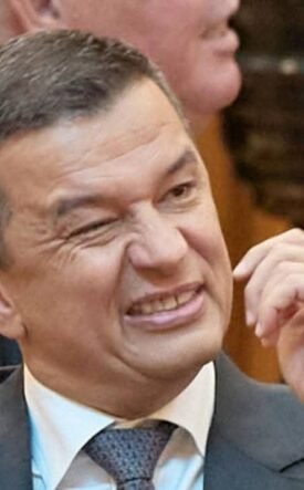 Man in a suit grimaces while rubbing his cheek at a meeting conference room scene.