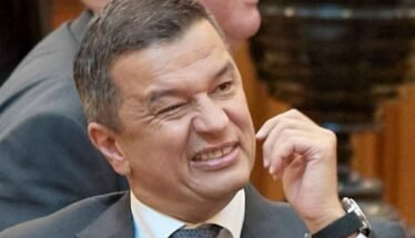 Man in a suit grimaces while rubbing his cheek at a meeting conference room scene.