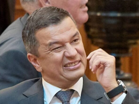Man in a suit grimaces while rubbing his cheek at a meeting conference room scene.