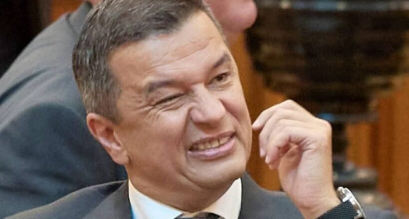 Man in a suit grimaces while rubbing his cheek at a meeting conference room scene.