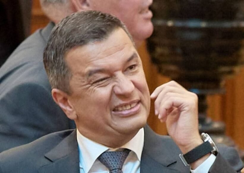 Man in a suit grimaces while rubbing his cheek at a meeting conference room scene.