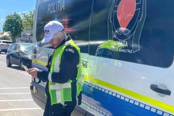 Policeman in a bright vest stands beside a blue-and-white police van, looking at a handheld device.