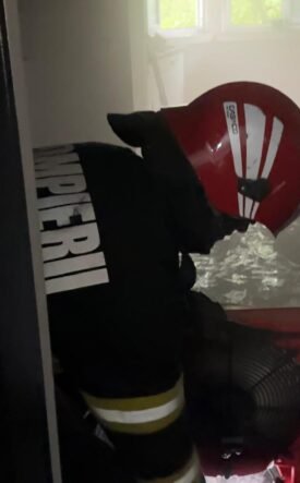 Firefighter in a red helmet and turnout gear crouches in a water-filled hallway during an emergency scene.