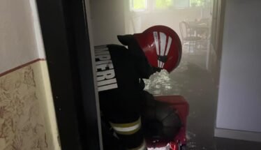 Firefighter in a red helmet and turnout gear crouches in a water-filled hallway during an emergency scene.