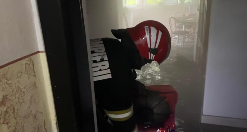 Firefighter in a red helmet and turnout gear crouches in a water-filled hallway during an emergency scene.
