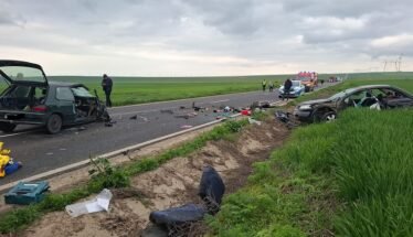 Two damaged cars on a rural highway after a crash, debris scattered across the road, responders and an ambulance in the distance with green fields around.