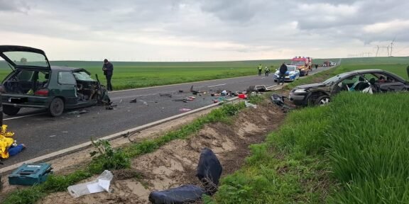 Two damaged cars on a rural highway after a crash, debris scattered across the road, responders and an ambulance in the distance with green fields around.