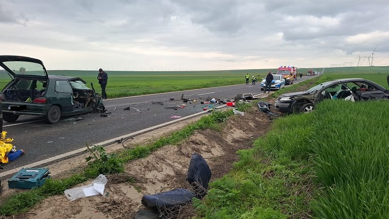 Two damaged cars on a rural highway after a crash, debris scattered across the road, responders and an ambulance in the distance with green fields around.