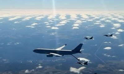 Large military tanker aircraft refuels a fighter jet in mid-air, with other jets nearby over a partly cloudy sky.