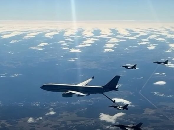 Large military tanker aircraft refuels a fighter jet in mid-air, with other jets nearby over a partly cloudy sky.