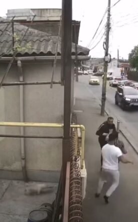 Group of men in a street confrontation on a sidewalk as cars drive by on a city road.