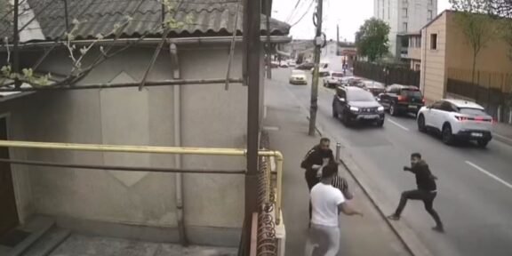 Group of men in a street confrontation on a sidewalk as cars drive by on a city road.
