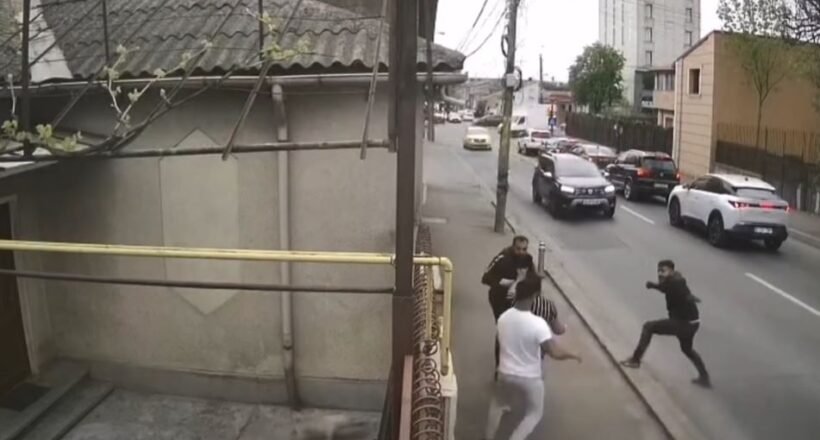 Group of men in a street confrontation on a sidewalk as cars drive by on a city road.