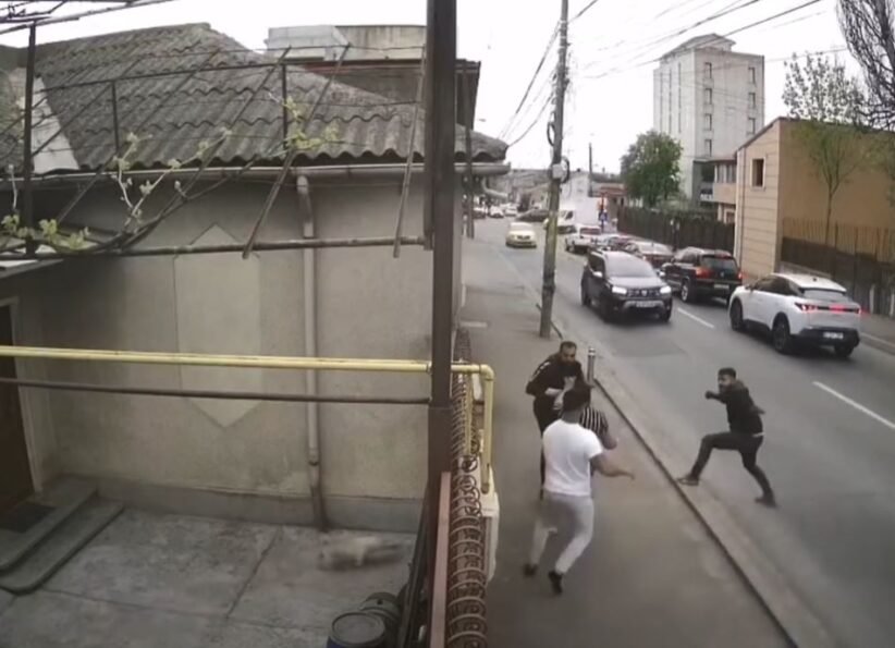 Group of men in a street confrontation on a sidewalk as cars drive by on a city road.