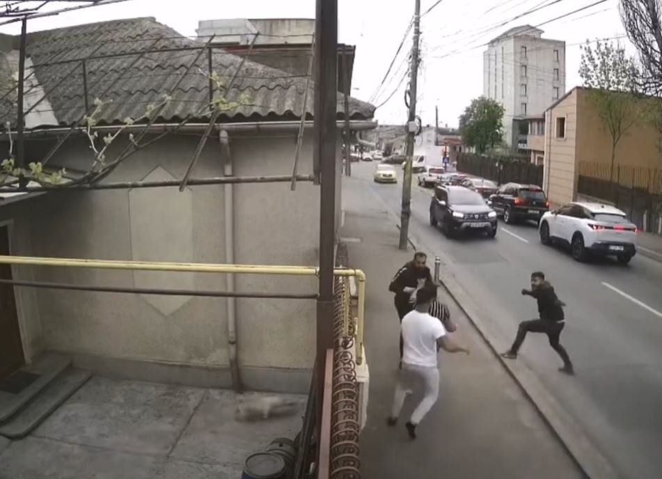 Group of men in a street confrontation on a sidewalk as cars drive by on a city road.