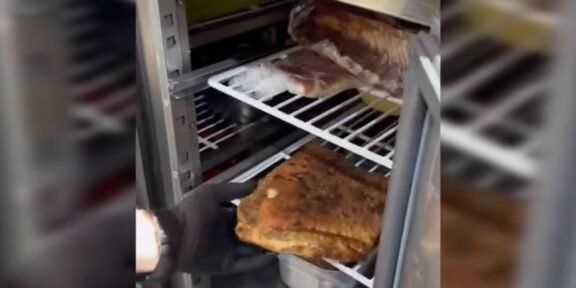 Person in a black glove lifts a large browned roast from a metal smoker/oven rack.