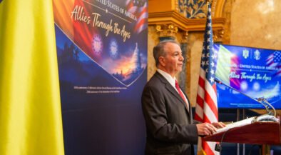 Man in a suit speaking at a podium during an Allied history event, with Romanian and American flags nearby.