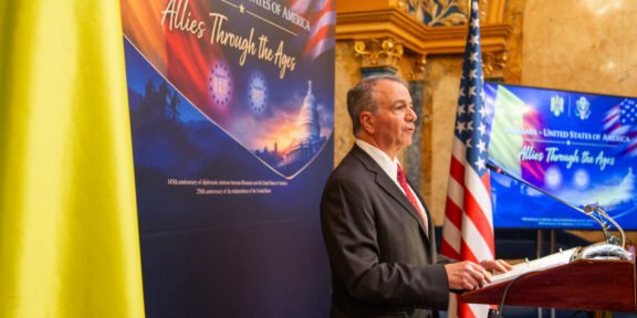Man in a suit speaking at a podium during an Allied history event, with Romanian and American flags nearby.
