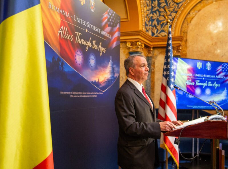 Man in a suit speaking at a podium during an Allied history event, with Romanian and American flags nearby.