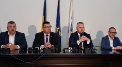 Four men in suits sit at a press conference table with microphones, flags behind them.