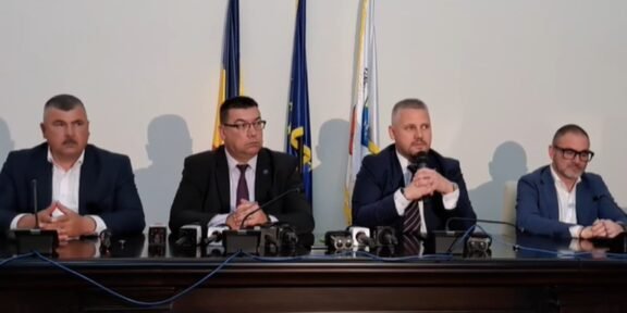 Four men in suits sit at a press conference table with microphones, flags behind them.