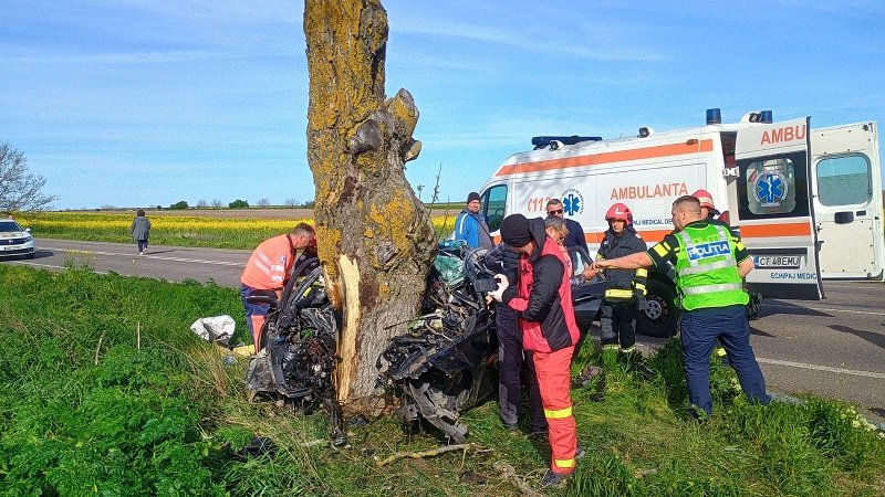 Emergency responders work at a car crash scene by a damaged vehicle against a tree; an ambulance is parked on a rural road.