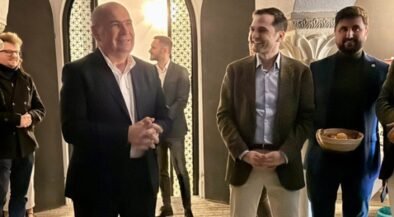 Group of people in a stone-arched hallway; a bald man in a dark suit speaks as a smiling man in a brown blazer stands nearby.