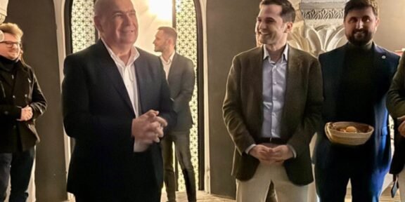 Group of people in a stone-arched hallway; a bald man in a dark suit speaks as a smiling man in a brown blazer stands nearby.