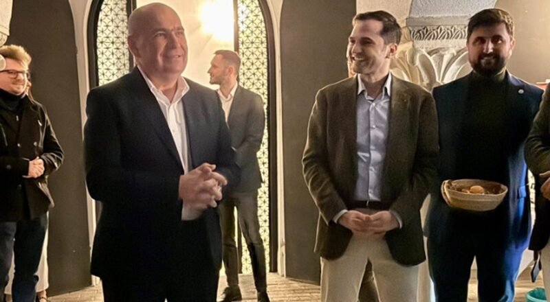 Group of people in a stone-arched hallway; a bald man in a dark suit speaks as a smiling man in a brown blazer stands nearby.