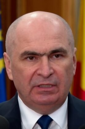 Bald man in a suit speaking at a press conference with microphones in front and flags in the background.