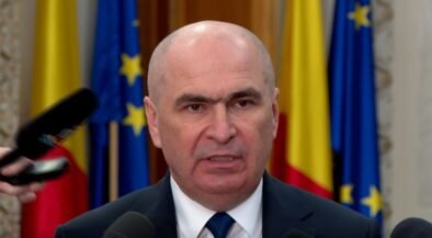 Bald man in a suit speaking at a press conference with microphones in front and flags in the background.