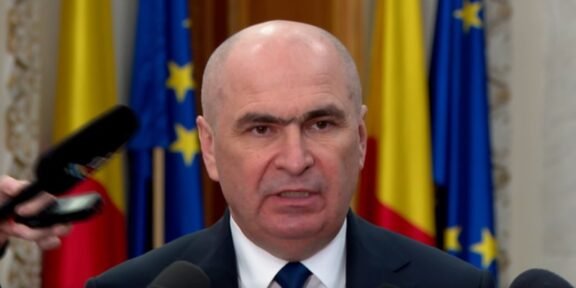 Bald man in a suit speaking at a press conference with microphones in front and flags in the background.